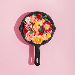 Creative concept with colorful roses, spring flowers and green leaves in frying pan on pastel pink...