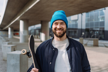 Trendy cool men portrait took to the camera, holding skate board and wearing cap. Skateboard spot. 