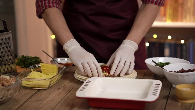 Cook in form prepares enchilada and roll up ingredients in tortilla in plate in kitchen restaurant. Man in gloves puts finished dish on baking tray against the background of mexican restaurant kitchen