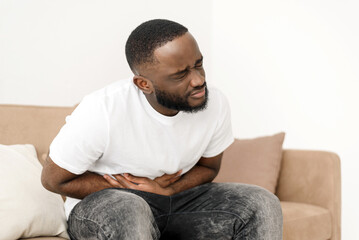 Black man with abdominal pain suffering from painful abdominal cramp in stomach sitting on couch at...