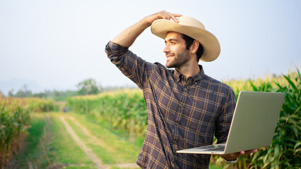Handsome Middle Eastern farmer holding a laptop in a corn field in wide-brimmed millet hat smiles...