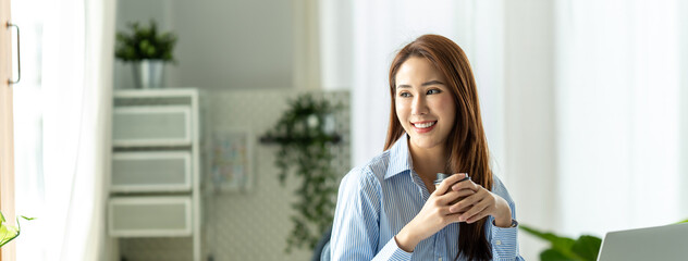 Asian business girl in her workstation at holding coffee cup while smile. Banner size.