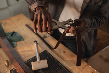 Midsection of african american young craftsman making hole on leather belt with punch plier