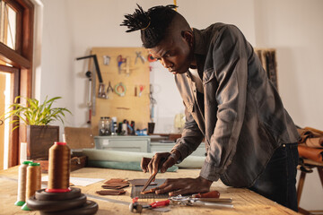 African american young craftsman measuring leather with scratch compass on cutting mat at workbench