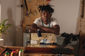 Portrait of african american young craftsman with arms crossed on sewing machine in leather workshop