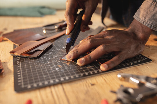 Cropped hands of african american young craftsman cutting leather with blade on cutting mat