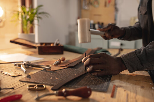 Midsection of african american young craftsman making hole on leather belt with hammer and equipment