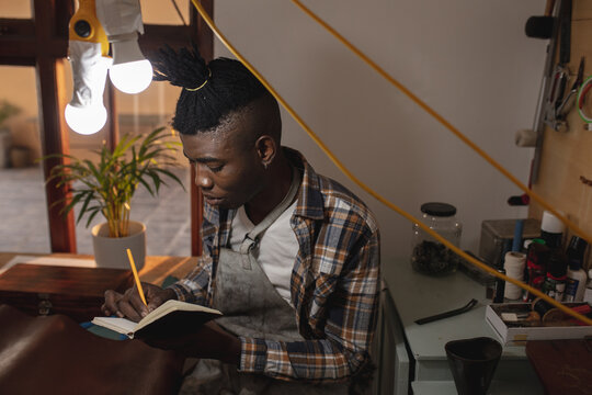 African american young craftsman writing on book while sitting in leather workshop