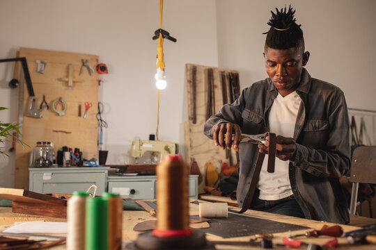 African american young craftsman punching leather belt with punch plier while working in workshop