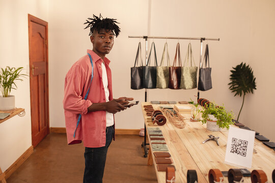 Portrait of confident african american male owner with smart phone standing in small leather store