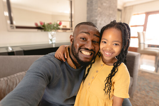 Portrait of smiling african american father and daughter taking selfie together sitting home