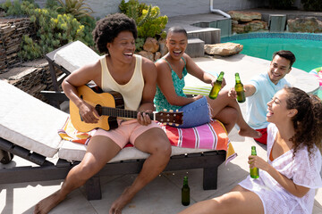 Happy multiracial friends toasting beer bottles while sitting by man playing guitar at poolside