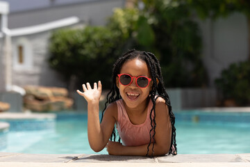 Portrait of happy african american girl wearing sunglasses leaning at poolside waving on sunny day