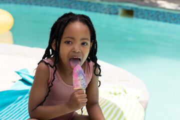 Portrait of cute african american girl eating ice cream while sitting at poolside on sunny day
