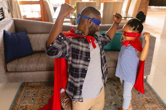 African american father and daughter wearing superhero costumes flexing muscles in living room