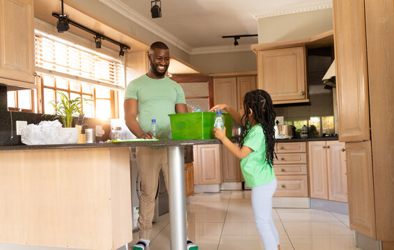Happy african american father and daughter collecting plastic bottles in recycling container at home
