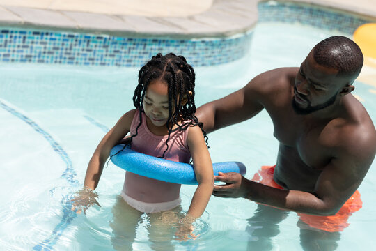 Shirtless african american father helping daughter with swimming float in pool on sunny day