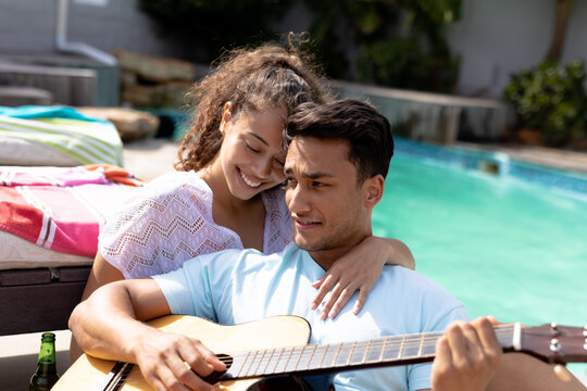 Happy multiracial couple enjoying sunny day at poolside while sitting with guitar
