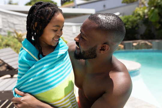 Smiling african american man wrapping daughter in blue towel at poolside on sunny day