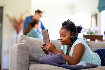 African american girl using tablet pc on sofa while caucasian female parents in background at home