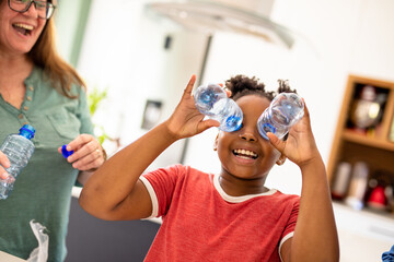 Caucasian mature mother looking at playful african american girl holding plastic bottles over eyes