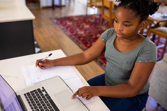 African american girl using laptop while writing on book during online school at home