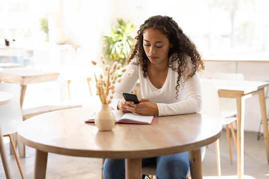 Young african american woman using smartphone while sitting with diary at table in coffee shop