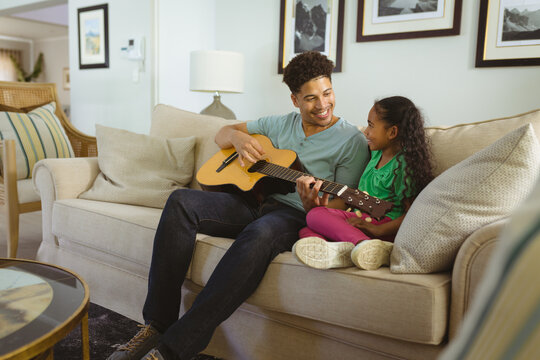 Happy multiracial father playing guitar while sitting together with daughter on sofa in living room