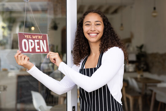 Happy young african american female barista showing open sign hanging on cafe door