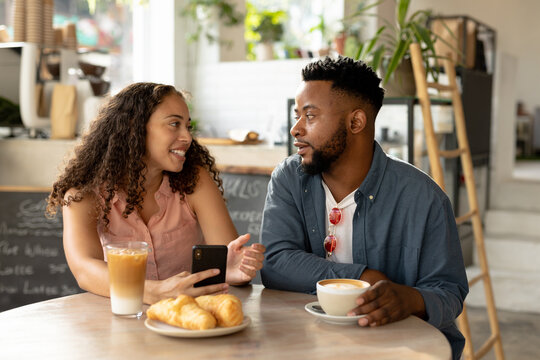 Happy african american couple talking while sitting with food and drink and smartphone at cafe