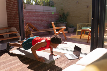 African american young man doing push-ups on exercise mat by laptop at home