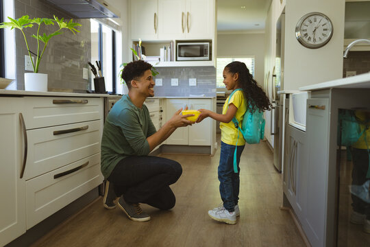 Biracial man giving lunch box to daughter while crouching in kitchen