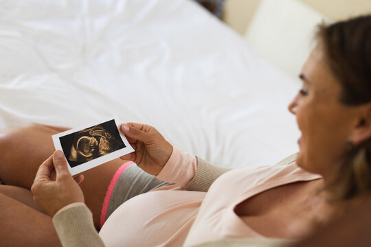 Caucasian pregnant woman smiling while holding ultrasound image of her baby at home
