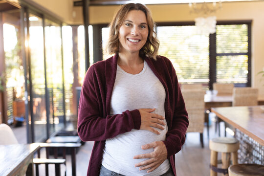 Portrait of caucasian pregnant woman touching her belly and smiling at home
