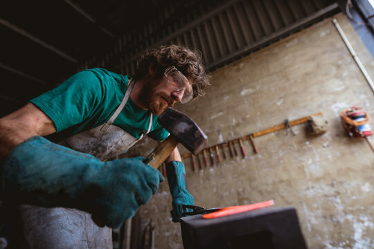 Bearded caucasian blacksmith in protective workwear forging with hammer on anvil in industry