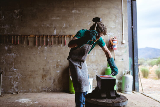 Caucasian blacksmith in protective gloves forging with hammer on anvil in manufacturing industry