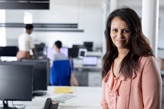 Portrait of confident businesswoman at desk in creative office while colleagues in background