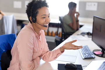 Smiling biracial female customer representative gesturing while talking through headset in office