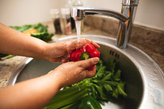 Cropped image of woman washing red bell pepper below running faucet at sink in kitchen