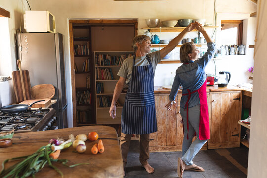Mature caucasian couple dancing while cooking food together in kitchen at home