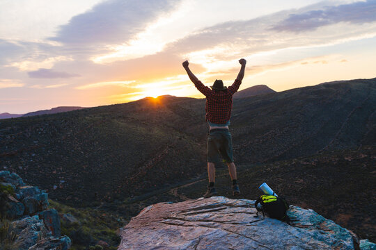 Excited young male caucasian hiker jumping from joy by backpack on mountain peak at sunset