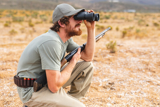 Side view of young caucasian male hunter crouching with rifle while looking through binoculars