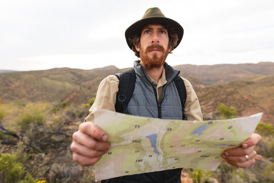 Bearded young male caucasian wanderer looking away while standing with map in wilderness