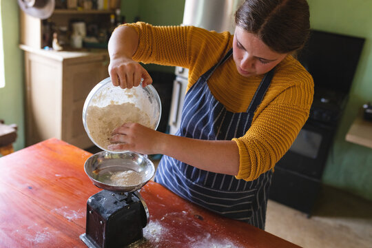 Young woman pouring flour from container on weight scale at table in kitchen