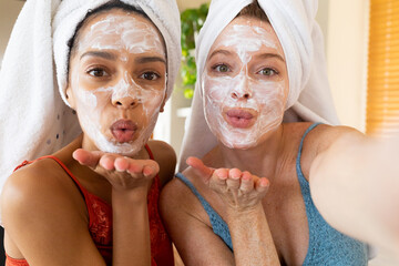 Portrait of female friends with facial masks and towels wrapped on hair blowing kisses at home