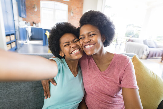 Cheerful african american woman taking selfie with teenage daughter while sitting on sofa at home