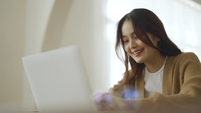 Smiling asian young woman working on laptop at home office. Young asian student using computer remote studying, virtual training, e-learning, watching online education webinar at house
