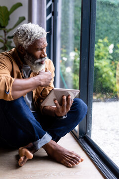 Thoughtful african american man looking through window sitting on floor with tablet pc at home