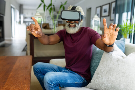 Senior african american man sitting on sofa gesturing while watching virtual reality simulator - Powered by Adobe