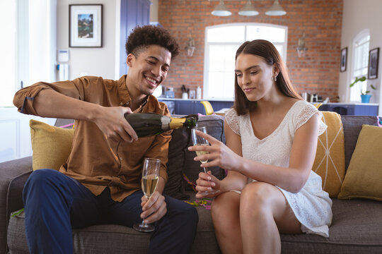 Happy young biracial man pouring champagne in flute for girlfriend at home